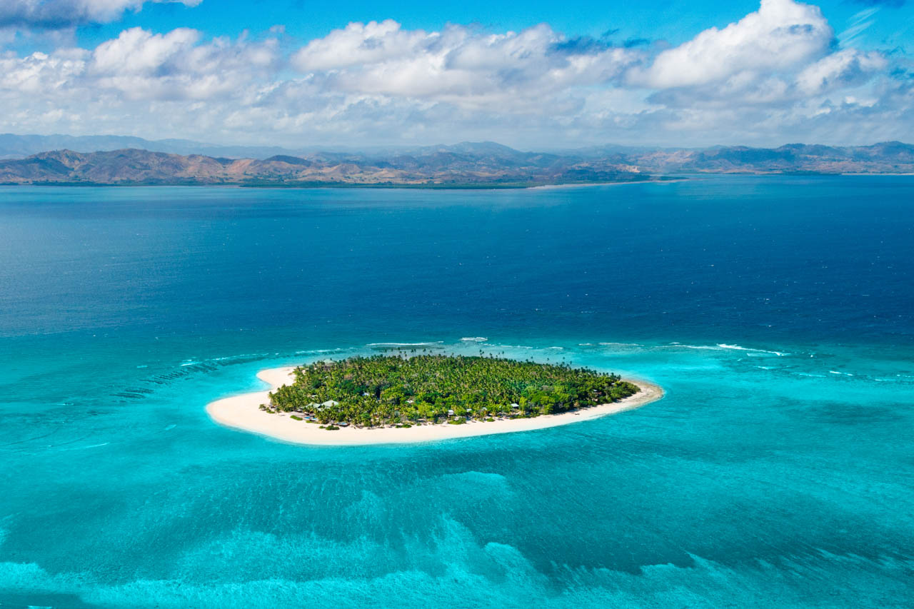 Drone photo of a small palm-covered island with white sand beaches and azure waters in the Yasawa group, Fiji.