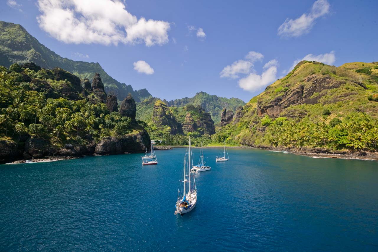 Sailboats anchored in a remote bay surrounded by rugged cliffs and dense vegetation in the Marquesas Islands.