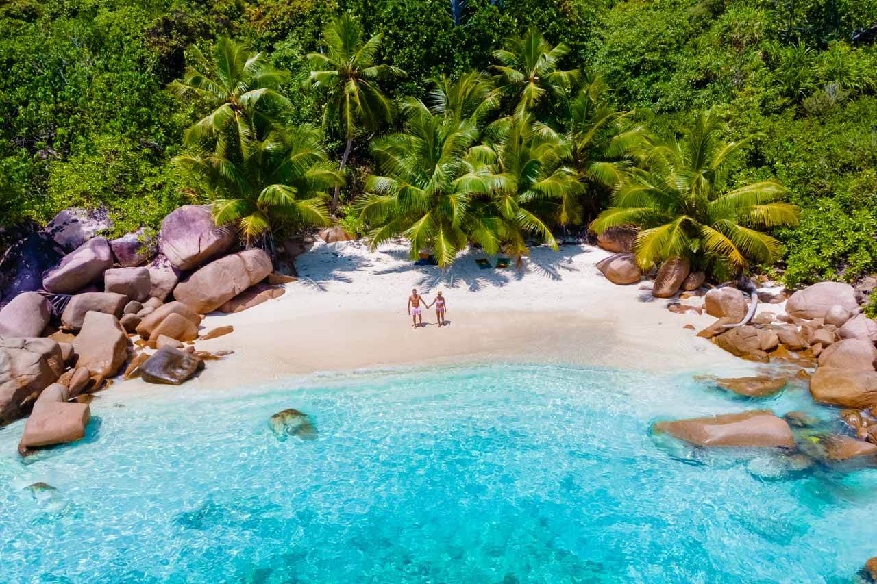 Couple standing on a small private beach with palm trees and turquoise water in Seychelles.